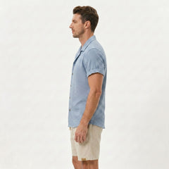 Side view of man in light blue textured Cuban shirt and linen drawstring shorts on plain background