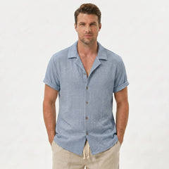 Three quarter view of man in light blue textured Cuban shirt and drawstring shorts on plain background