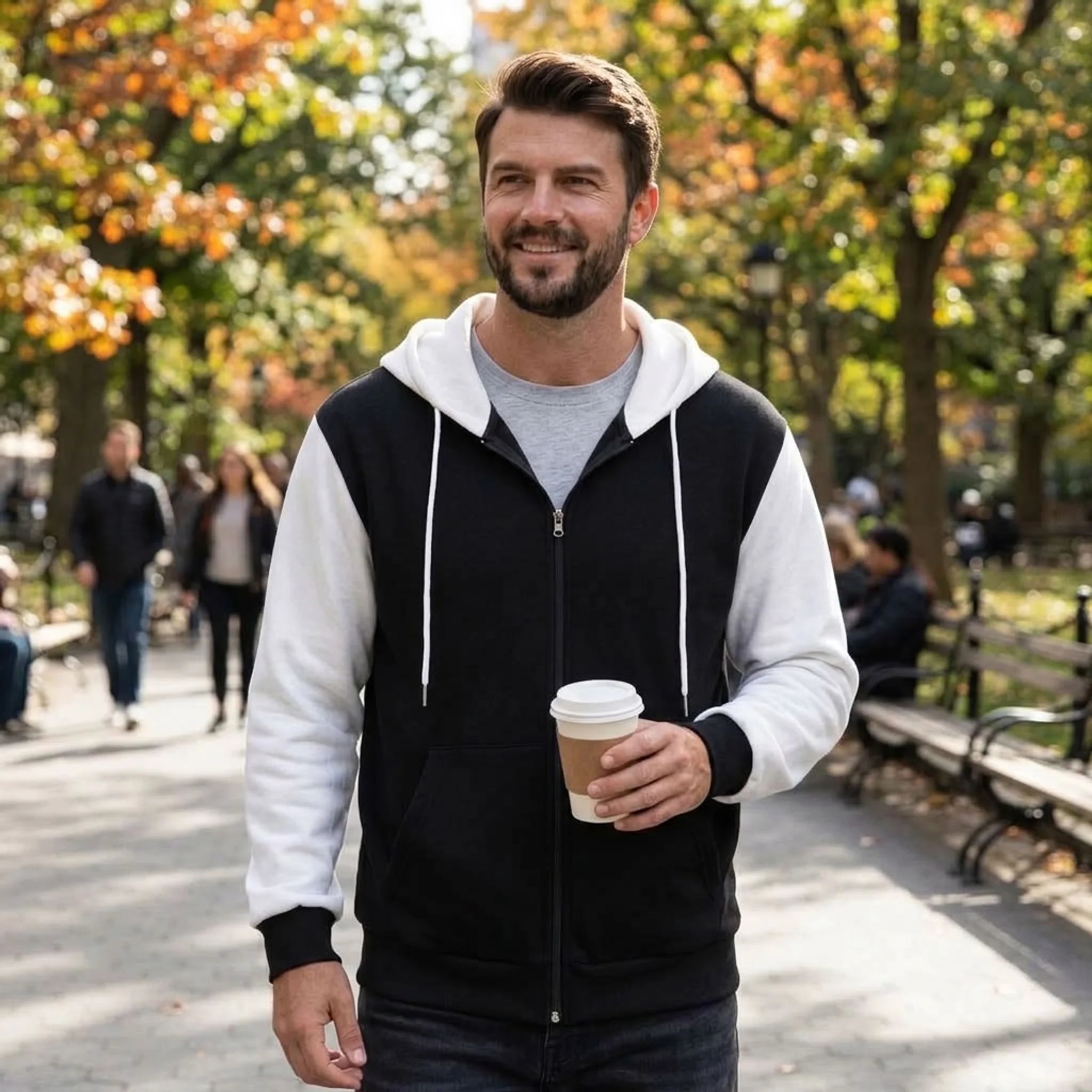 Man holding a coffee wearing black and white zip-up hoodie in an autumn park