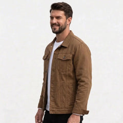 A man wearing a coffee utility-style trucker jacket over a white t-shirt and black pants, standing against a light gray textured background.