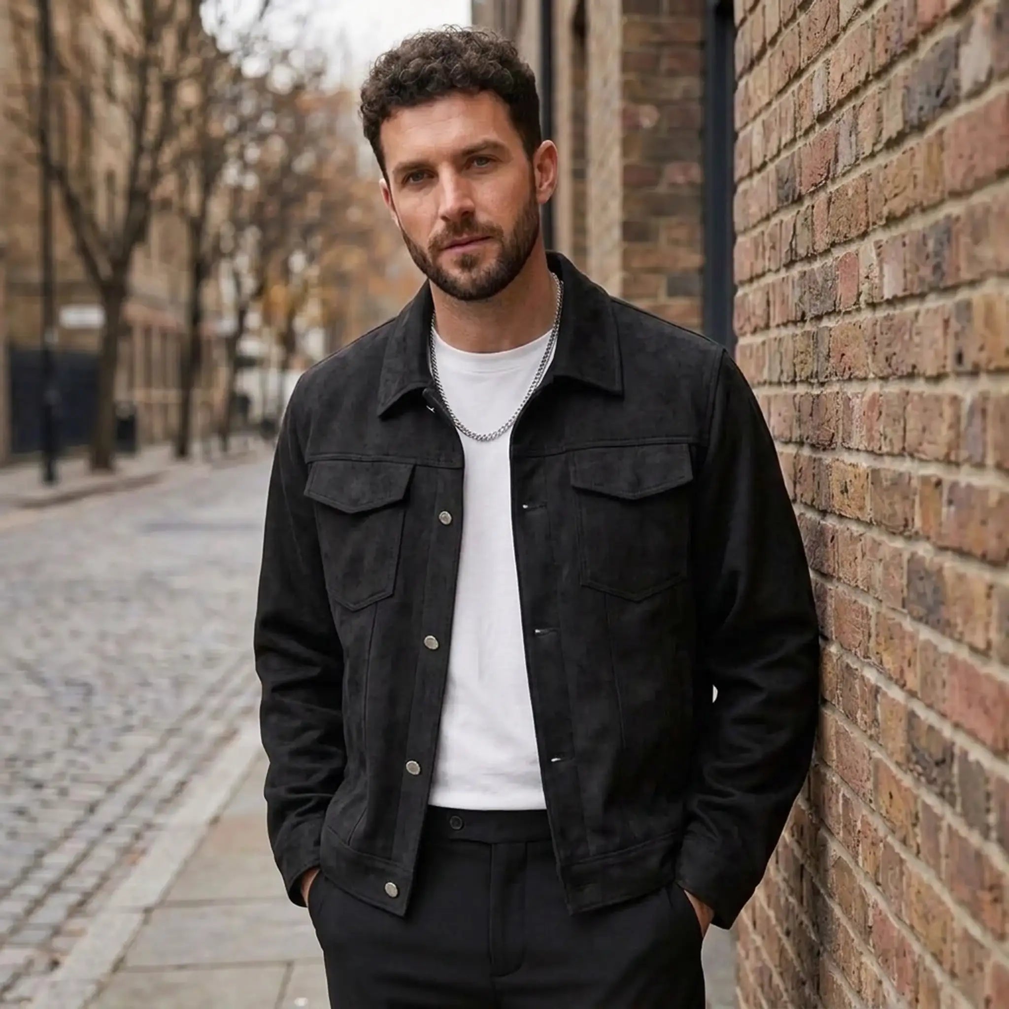 Man in a black suede-look trucker jacket over a white tee, leaning against a brick wall on a city street