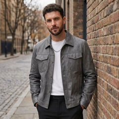 Man in a gray suede-look trucker jacket over a white tee, leaning against a brick wall on a city street
