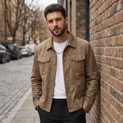 Man in a khaki suede-look trucker jacket over a white tee, standing against a brick wall on a city street