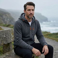 MannerCo dark grey fleece hoodie, man seated on a stone wall by the sea