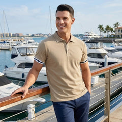 Man in khaki polo shirt with striped cuffs leaning on marina railing with yachts in background