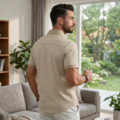 Man in khaki waffle knit polo shirt gazes out window holding coffee cup in bright modern living room