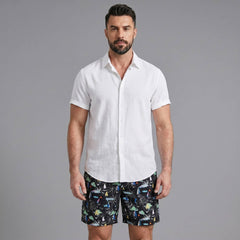 Man wearing a white linen rayon button up shirt and black palm tree print swim shorts front view on plain background