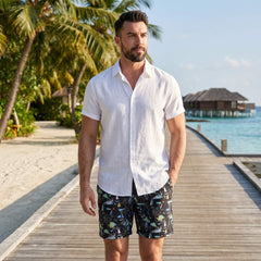 Man wearing white linen rayon button up shirt and black palm tree swim shorts in outdoor lifestyle setting