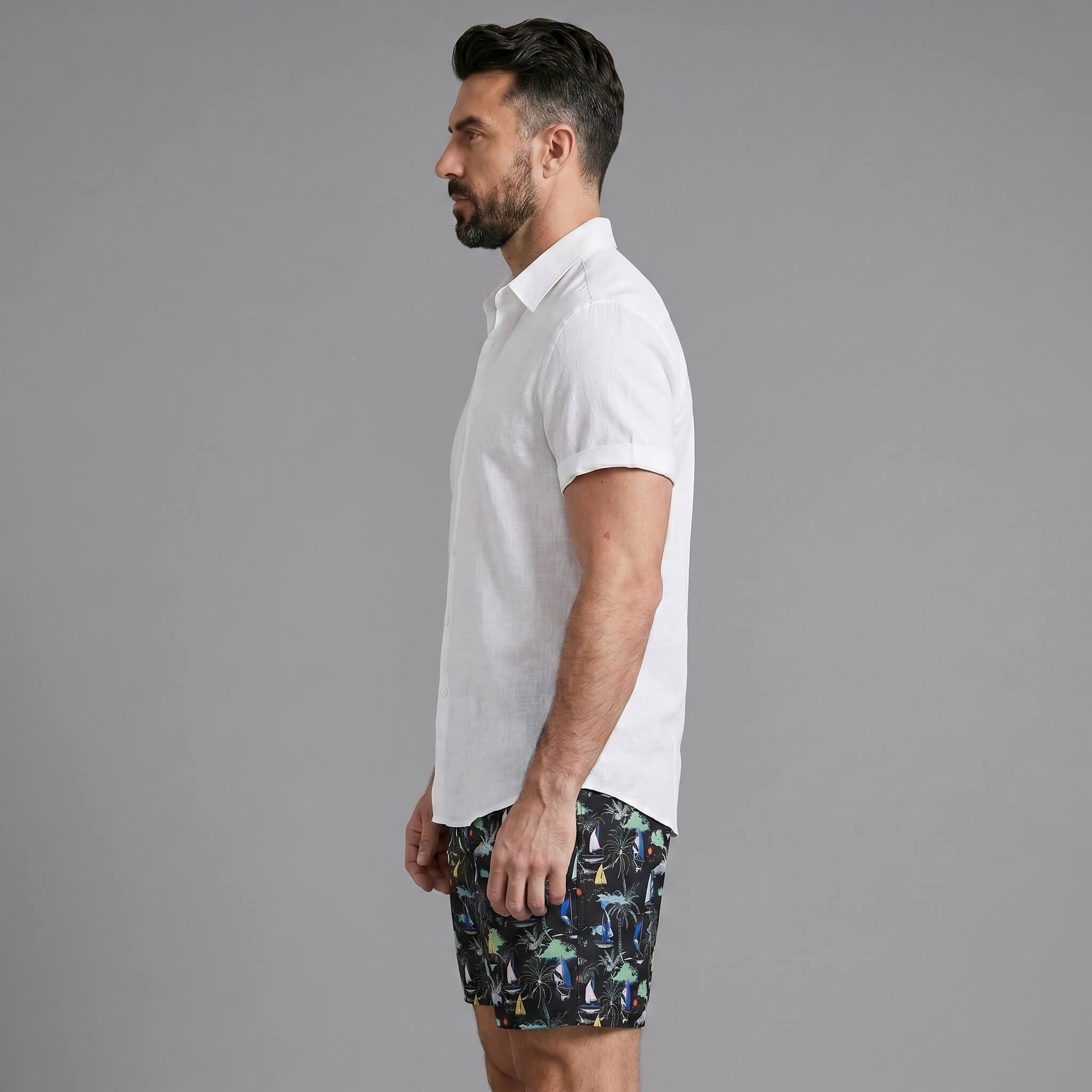 Side view of man in white linen rayon button up shirt and black palm tree print swim shorts