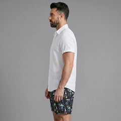 Side view of man in white linen rayon button up shirt and black palm tree print swim shorts