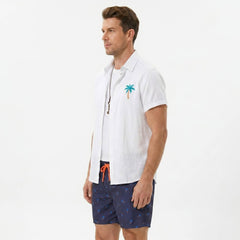 Side view of man in white palm tree print linen look shirt and swim shorts on plain background
